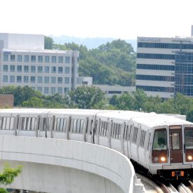 A photographic image of a light rail train traveling along a bridge with a city background.