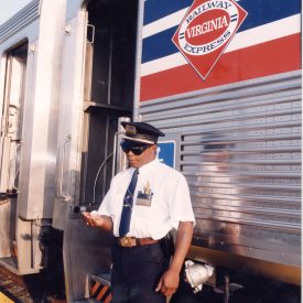 A photographic image of a Virginal Railway Express (VRE) conductor in front of a stationary passenger train.